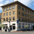 Pixelated static image of a multi-story building with light and dark brown facades, multiple windows, and a streetlamp in front under a blue sky.