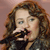 A pixelated close-up of a person with long brown hair holding a microphone, wearing a dark jacket over a light shirt against a muted pink background.