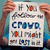 A person holds a white sign with text reading "IF YOU FOLLOW THE CROWD, YOU MIGHT BE LOST IN IT." The sign features blue and orange lettering against a light background.