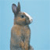 Pixelated image of a rabbit standing upright against a light blue background, with brown and gray fur tones and visible ear structure.