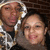 Two people in hooded jackets pose together against a brick wall background, with one person's head slightly tilted.