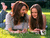 Two people recline on grassy ground; one wears a light-colored top and dark pants, the other a dark sleeveless top. Background shows greenery with yellow accents.