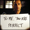 A person holds a white sign with handwritten text against a dark background with windows and framed items.