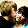 Two people in close physical proximity, one with light brown hair kissing the other on the cheek, both wearing dark and light blue tops.