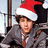 A person wearing a red and white Santa hat and gray jacket with a brown strap, standing next to a white background with red text reading 'merry christmas'.