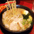 A bowl of ramen with noodles, green vegetables, and a round white topping on red background.