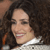 A pixelated close-up of a person with dark curly hair wearing a white top, smiling with visible teeth against a dark background.