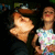 Two people in a close-up pose with one person leaning toward the other; background is dark and indistinct.