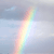 A vertical rainbow arc spans a soft blue-gray sky with faint cloud patches, rendered in pixelated style with pastel hues transitioning from pink to green.