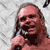 Pixelated static image of a person with long dark hair holding a microphone against a light gray background, rendered in low-resolution 50x50 pixels with visible dithering and color banding.