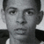 Static grayscale image of a person's face with short dark hair, wearing a dark jacket over a light shirt, cropped to show shoulders and upper chest.