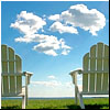 Two light green Adirondack chairs face a blue sky with white clouds over a grassy field and distant ocean horizon.
