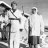 Black-and-white pixelated image of two figures in white shirts and dark pants standing side-by-side with arms raised, set against a gradient background.