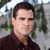 Pixelated portrait of a person with short dark hair wearing a dark collared shirt against a blurred outdoor background with green foliage and light sky.