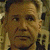 Pixelated close-up of a person's face with light brown hair, wearing a dark shirt, against a blurred background with green and brown tones.