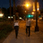 Two figures walk along a dimly lit path under glowing streetlights, with blurred trees and distant lights in the background.