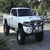 Static pixelated image of a light-colored pickup truck with dark wheels and grille, positioned on a road against blurred greenery background.