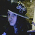 A person wearing a black hat with a blue and white checkered emblem, dark clothing, and a light-colored background with a blurred figure behind.