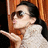 A person with long dark hair and sunglasses blows a kiss, wearing a white textured sweater against an orange-brown background.