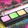 A close-up of a makeup palette with four square pans of colored powder in pink, green, yellow, and silver, set against a glittery multicolored background.