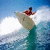 Animated surfing icon showing a person riding a wave on a surfboard across two sequential frames with motion blur and water spray effects.
