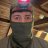 Pixelated figure wearing dark mask covering lower face, pink headband with blue section, and dark clothing against blurred background.