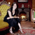 A pixelated figure in a dark dress stands in a room with warm lighting, holding a white object near a fireplace.