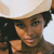 A pixelated close-up of a person wearing a light-colored wide-brimmed hat and dark clothing, with a neutral expression and visible shoulder.