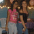 Two women stand side-by-side in a dimly lit indoor setting with string lights in the background; one wears a pink graphic top and light pants, the other a black off-shoulder top and dark pants.