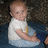 A baby in a white shirt and light blue pants sits on a dark surface with arms crossed, head slightly turned left.