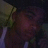 Pixelated static image with dark background, indistinct figure in muted green and brown tones holding a light-colored object near the face.