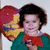 A person with short dark hair wears a green top and red bow, holding an object against a heart-shaped backdrop.