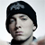 Pixelated close-up of a person wearing a black beanie with a gray pattern, light skin, and a dark collar visible at the neck.