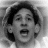 Black-and-white pixelated image of a person with short dark hair, mouth open in a shout or yell, visible teeth, and a blurred background.