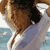Pixelated static image of a person in swimwear on a beach with water and sky background, rendered as a tiny buddy icon.