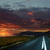 A road stretches toward a sunset with orange and purple sky, mountains in the background, and white lane markings on asphalt.