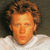 Pixelated close-up of a person with light brown curly hair and a white shirt against a dark background.