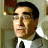 Pixelated image of a person with short dark hair wearing a white shirt and dark tie, standing against a blurred background with indistinct shapes and muted colors.