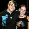 Two people stand side-by-side against a dark background; the right person's shirt displays 'Memories' text in yellow pixelated letters.