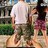 Pixelated static image of two figures walking outdoors, one in white shirt and dark pants, the other in purple top and dark pants, with greenery and buildings in background.