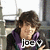 Pixelated image of a person with brown hair wearing a purple and black shirt with white text, seated in an indoor setting with blurred background elements.