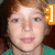 A pixelated close-up of a person with light brown hair wearing a green top and dark necklace, with an orange and white icon in the top right corner.