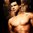 A shirtless muscular man with dark hair stands against a black background, his right arm raised and left hand near his chest.