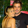 Two people pose closely together against a green foliage background with pink accents, one in a black shirt with a graphic design, the other in a brown strapless top.