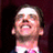 A pixelated image of a person with light skin and dark hair, wearing a white shirt and pink tie against a blue background with text.