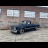 A dark blue pickup truck is parked in front of a brick building with three white garage doors, viewed from a low angle on a light-colored concrete surface.