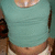 A person wearing a teal crop top and light-colored shorts, with a visible belly button and a blurred background featuring dark shapes.