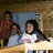 A pixelated image of a girl with long dark hair in a white shirt with pink accents, seated at a table with others in a dimly lit room, with text overlays changing across frames.