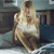 Pixelated image of a person with long blonde hair wearing a white sleeveless top and brown pants, seated in a room with dark furniture and blue flooring.