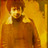 Pixelated figure with reddish-brown hair and a brown top, hand near face, against warm yellow-orange background.